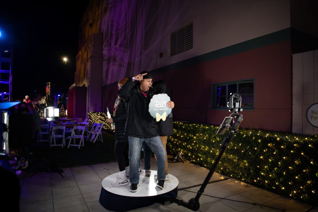 Three guests standing on circular platform with cameras circling them to take a 360-degree photo. The guest facing the camera is wearing a black coat, blue jeans and is holding a D23 Christmas Wreath photo prop. The guests behind that guest are getting ready for the camera to come their way.