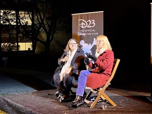 Rebecca Cline and Mary Walsh sitting on stage presenting. Cline is wearing a black coat, white sweater, and black pants. Walsh is to her right wearing a red coat and blue jeans.