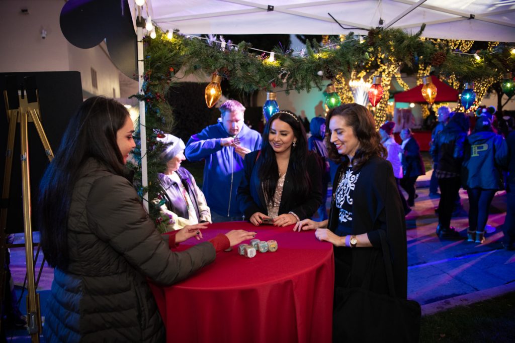 Two guests playing Jingle Jangle at the D23 Booth with cast member. The cast member is wearing a black winter coat standing behind a red table explaining how to play the game. The table has six large dice sitting on it. The guest on the left is wearing a black coat and gold dress. The guest on the right is wearing a black coat and black dress.