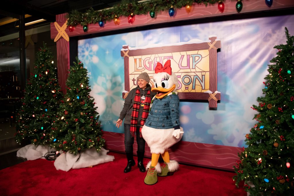Guest posing with Daisy in front of Light Up The Season photo backdrop. The backdrop is blue and snowy with the Light Up The Season logo of stained glass in yellow, blue and pink framed by wood in the center of the backdrop. Daisy is wearing a red bow and a blue winter coat. The guest has their arm around Daisy and is wearing a gray beanie, red checkered scarf, gray jacket and jeans.
