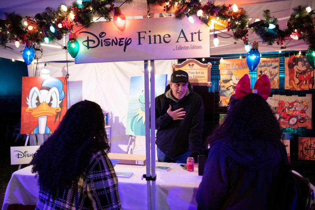 Guests shopping at the Disney Fine Art Booth. The cast member is wearing a black coat and black baseball bat. The cast member is talking to guests from behind the booth. The booth has Disney Fine Art on a sign at the top. At the back of the booth are a variety of art pieces and a large painting of Donald Duck.