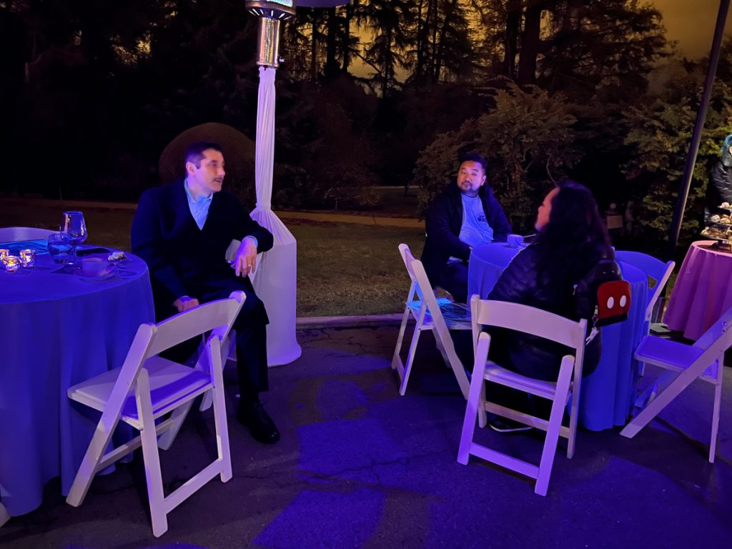Three guests sitting at two different tables talking. The guest on the right is wearing a blue shirt, black coat, and black pants. The guest on the left is wearing a blue shirt and black coat. The guest in front is wearing a black coat.