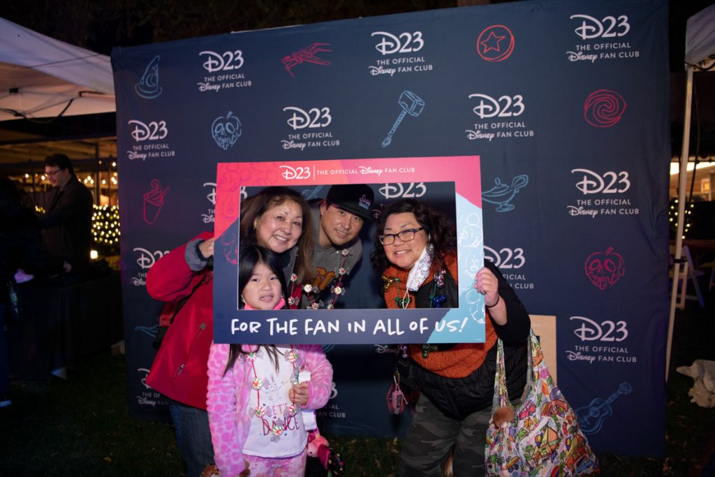 Four guests standing in front of the D23 logo photo backdrop holding a pink and blue frame cutout reading D23: The Official Disney Fan Club for the fan in all of us photo prop. The fan on the left is wearing a pink coat and blue jeans. The guest in front is wearing a pink jacket, white shirt, and pink pants. The guest in the middle is wearing a black baseball cap and gray sweater. The guest to the right is wearing an orange shirt, black sweater, and jeans.