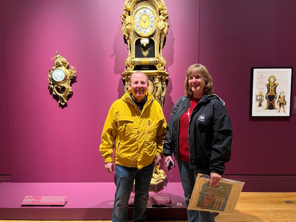 Two guests smiling in front of a gold clock. The guest on the left is wearing a yellow coat and blue jeans. The guest on the right is wearing a black coat, red sweater, and blue jeans.