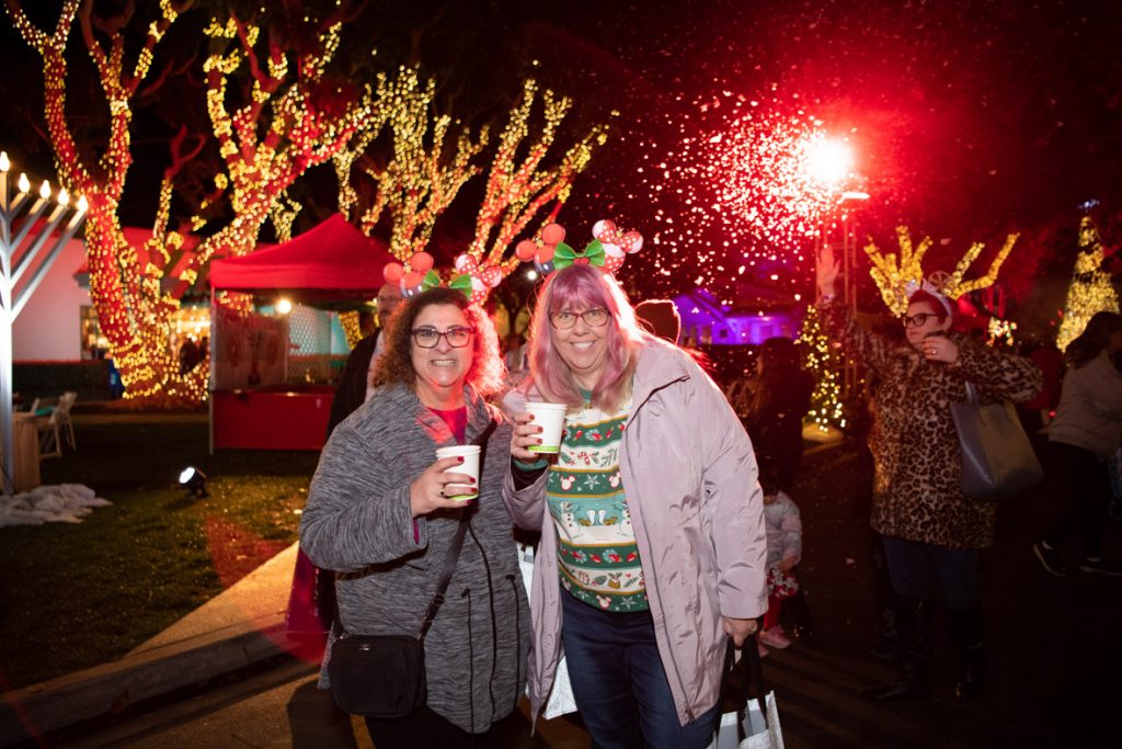 Two guests holding warm beverages in front of lit up trees. The guests are both wearing Minnie Mouse gingerbread ears. The guest on the left is wearing a long grey jacket. The guest on the right is wearing a green and white striped holiday Disney spirit jersey, blue jeans, and gray jacket.