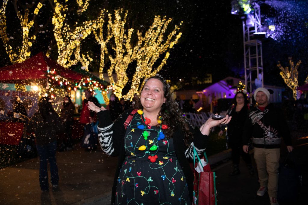 Guest smiling in front of snow falling and lit up trees. The guest is wearing a black dress with Mickey Christmas lights, black sweater, and lit up Christmas bulb necklace.