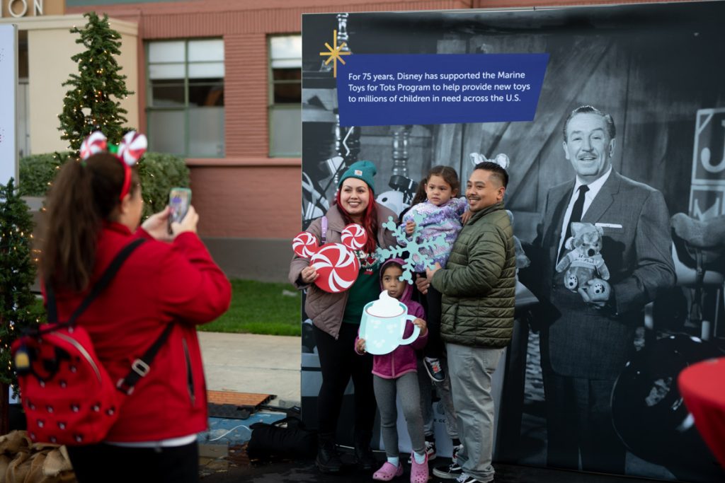 Guest taking a photo of a family in front of The Walt Disney Toys for Tots backdrop. The backdrop has a black and white photo of Walt Disney holding a stuffed bear. The guest taking the photo is wearing Candy Cane Minnie Mouse ears, a red jacket and black jeans. The guest in the photo on the left is holding a Peppermint Mickey photo prop. The guest is wearing a green hat, brown jacket and gray jeans. The guest in front is holding a blue hot coco mug photo prop. The guest is wearing a pink jacket, grey pants and pink shoes. The guest to the left is holding a guest who is holding a blue Mickey snowflake photo prop. The guest holding the prop is wearing a purple coat. The guest holding the guest is wearing a green coat and gray pants.