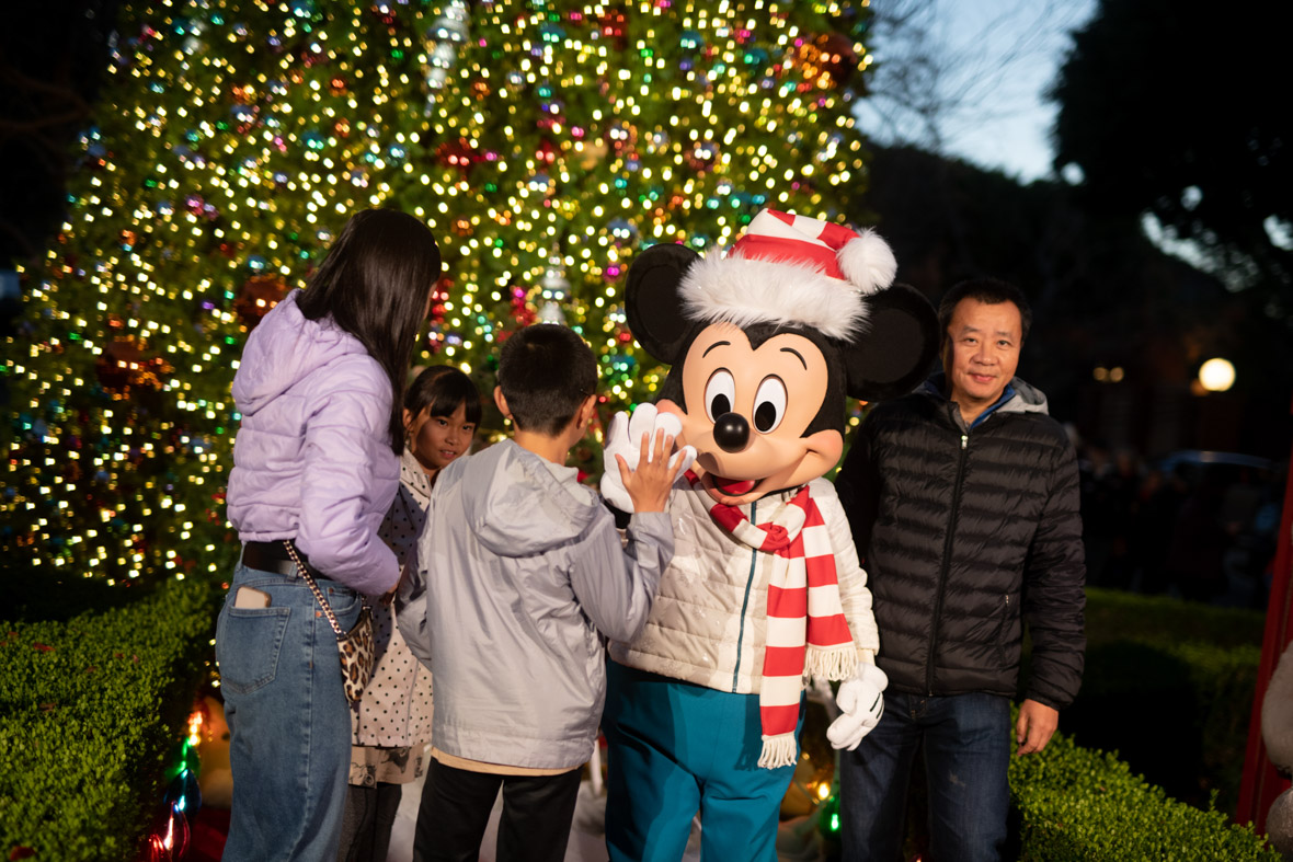 Guest high-fiving Mickey. Mickey is wearing a red and white striped Santa hat with a matching red and white striped scarf. He is wearing a white winter coat and teal pants. The guest high-fiving Mickey is wearing a silver coat and black pants. The guest to the left is wearing a purple jacket and blue jeans. The guest to the right of Mickey is wearing a black coat and blue jeans. They are all standing in front of a lit-up Christmas tree.