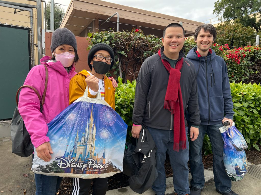 Four guests after shopping: The guest on the far right is wearing a grey beanie, pink raincoat, pink face mask, and jeans; this guest has their hand on the shoulder of the guest to their right. That guest is wearing a black beanie, black face mask, yellow raincoat, and black sweatpants. This guest is holding a blue Disney Parks bag. The guest to the right is wearing a red scarf, black jacket, and blue jeans. The guest to the far right is wearing a blue coat, blue jeans and is holding a blue Disney Parks bag.
