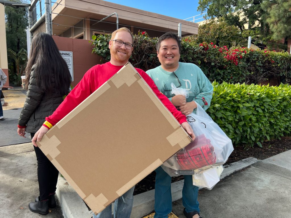 Two guests after shopping in Mickey’s of Glendale. The guest on the left is holding a large brown cardboard box, and is wearing a red sweater and jeans. The guest to the right is holding three clear Walt Disney Imagineering shopping bags, and is wearing a green Disneyland holiday sweater and jeans.