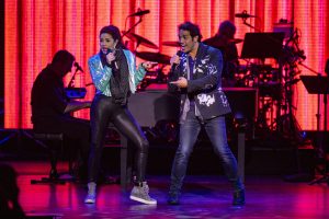 Broadway performers Arielle Jacobs and Adam Jacobs on stage, mid-song, in a promotional image for the 2023 DISNEY ON BROADWAY concert series at EPCOT, during the International Festival of the Arts. Arielle (on left) is wearing a shiny turquoise jacket, black leggings, and sparkly silver hi-top sneakers; Adam (on right) is wearing a black and white patterned suit jacket and grey scarf with dark jeans. A piano player and drummer are silhouetted behind them, with an orange and red lit-up background.