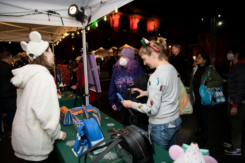 Guests looking and shopping at the Loungfly booth. The green table has a variety of different products, The employee is wearing a white jacket and white Mickey hat.