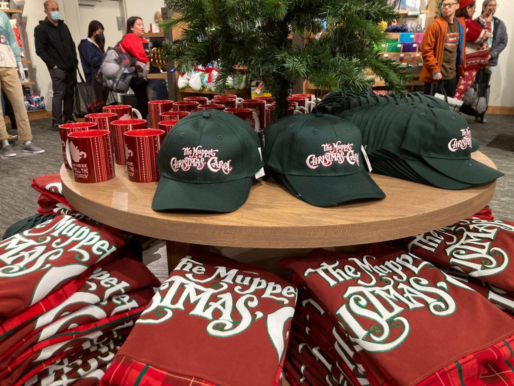 The Muppet Christmas Carol merchandise sitting on round wood tables. On the left are red coffee mugs with Gonzo on the. To the left are dark green baseball hats featuring The Muppet Christmas Carol logo. Below the hats are red spirit jerseys with The Muppet Christmas Carol written on the back in white.