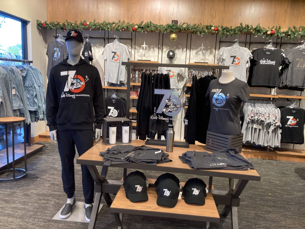 Walt Disney Imagineers 70th anniversary merchandise: On the left is a manikin with a black hat with the Walt Disney Imagineering 70th anniversary logo with sorcerer Mickey; it’s also wearing a black sweatshirt with Walt Disney Imagineering 70th anniversary logo and black sweatpants. On the wood table to the right of the manikin are grey shirts folded towards the front, black hats below, and white water bottles behind the shirts. On the right is a manikin bust displaying a grey t-shirt with the Walt Disney Imagineering 70th anniversary logo in silver.