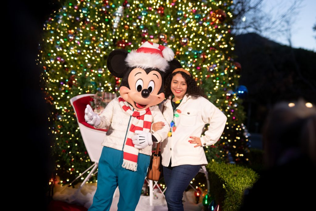 Guest posing with Mickey Mouse. Mickey is wearing a red and white striped Santa hat with a matching red and white striped scarf. He is wearing a white winter coat and teal pants. The guest to Mickey’s right is wearing a white jean jacket, blue jeans and has a light up Christmas bulb necklace.