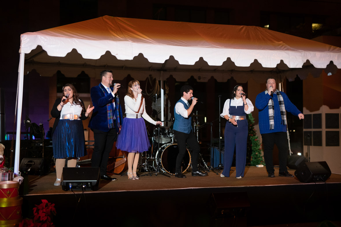 Mostly Kosher performing on stage. All singers are holding black microphones. The singer on the far left is wearing a black sweater, white shirt and blue skirt. The singer to the right is wearing a navy suit and blue scarf. The singer to the right is wearing a white sweater and blue skirt. The singer to the right is wearing a white shirt, blue vest and black slacks. The singer to the right is wearing a blue jumpsuit and white blouse. The singer to the far left is wearing a blue sweater, black slacks, and black and white checker scarf.