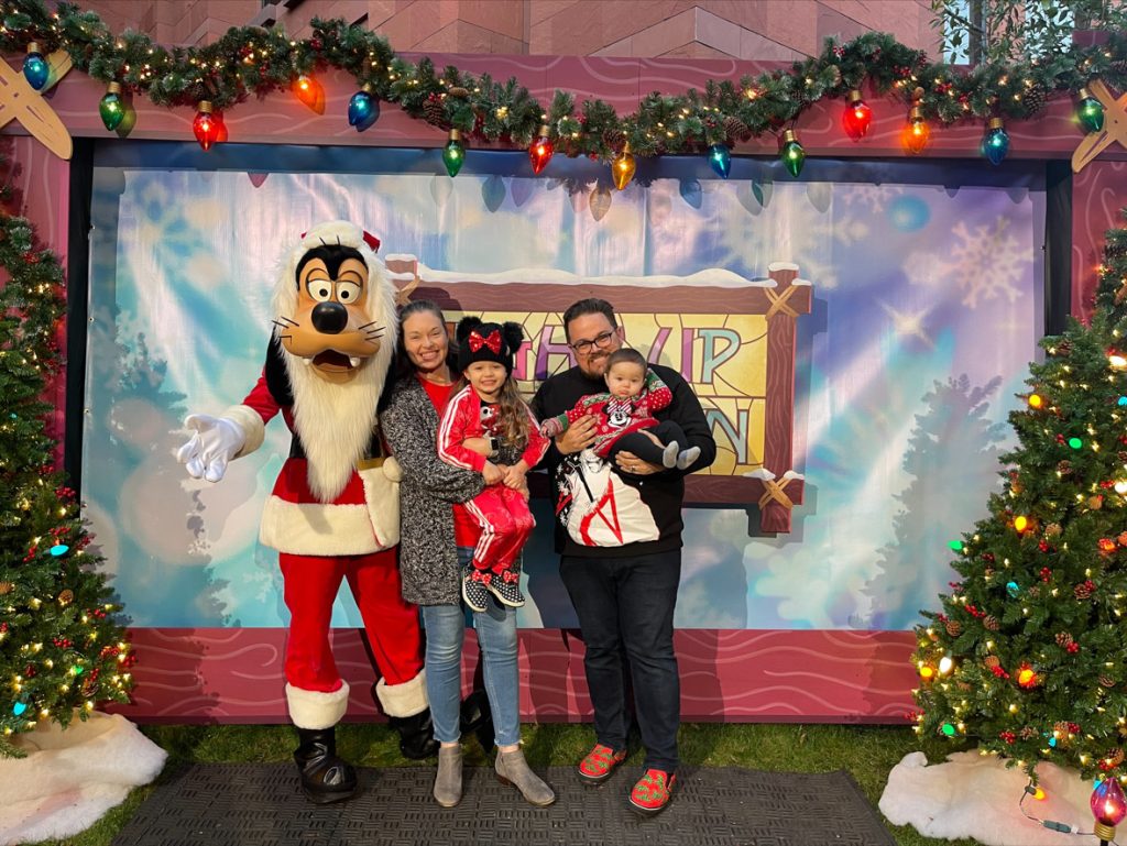 Four guests posing with Santa Goofy in front of a Light Up the Season photo backdrop. The backdrop is blue and snowy with the Light Up The Season logo of stained glass in yellow, blue and pink framed by wood in the center of the backdrop. Santa Goofy is on the far left side with his arm around the first guest. The guest is wearing a grey sweater, red shirt, blue jeans and grey boots. This guest is holding a guest who is wearing a Minnie Mouse hat, red sweatsuit and Minnie Mouse polka dot shoes. The guest to the right is wearing a black sweater, blue jeans, and red shoes. This guest is holding another guest who is wearing a Minnie Mouse holiday sweater and black pants.