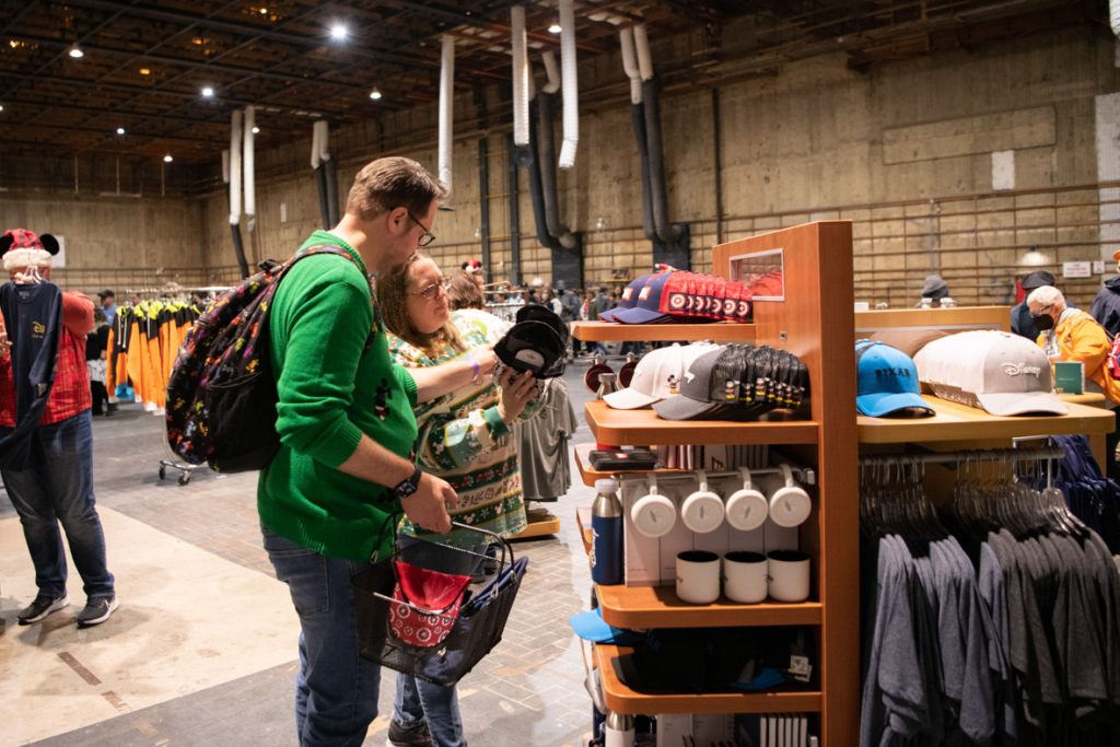 Guest shopping in the Employee Store. The guest on the left is wearing a green sweater and blue jeans. The guest on the right is wearing a Disney holiday spirit jersey in green and white stripes with jeans. They are both looking at a black baseball cap. Surrounding them are multiple displays of a variety of merchandise.