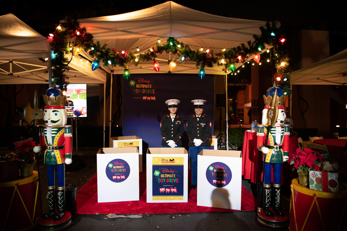 Two marines in uniform standing at the Toys for Tots booth behind the Disney Ultimate Toy Drive donation boxes.