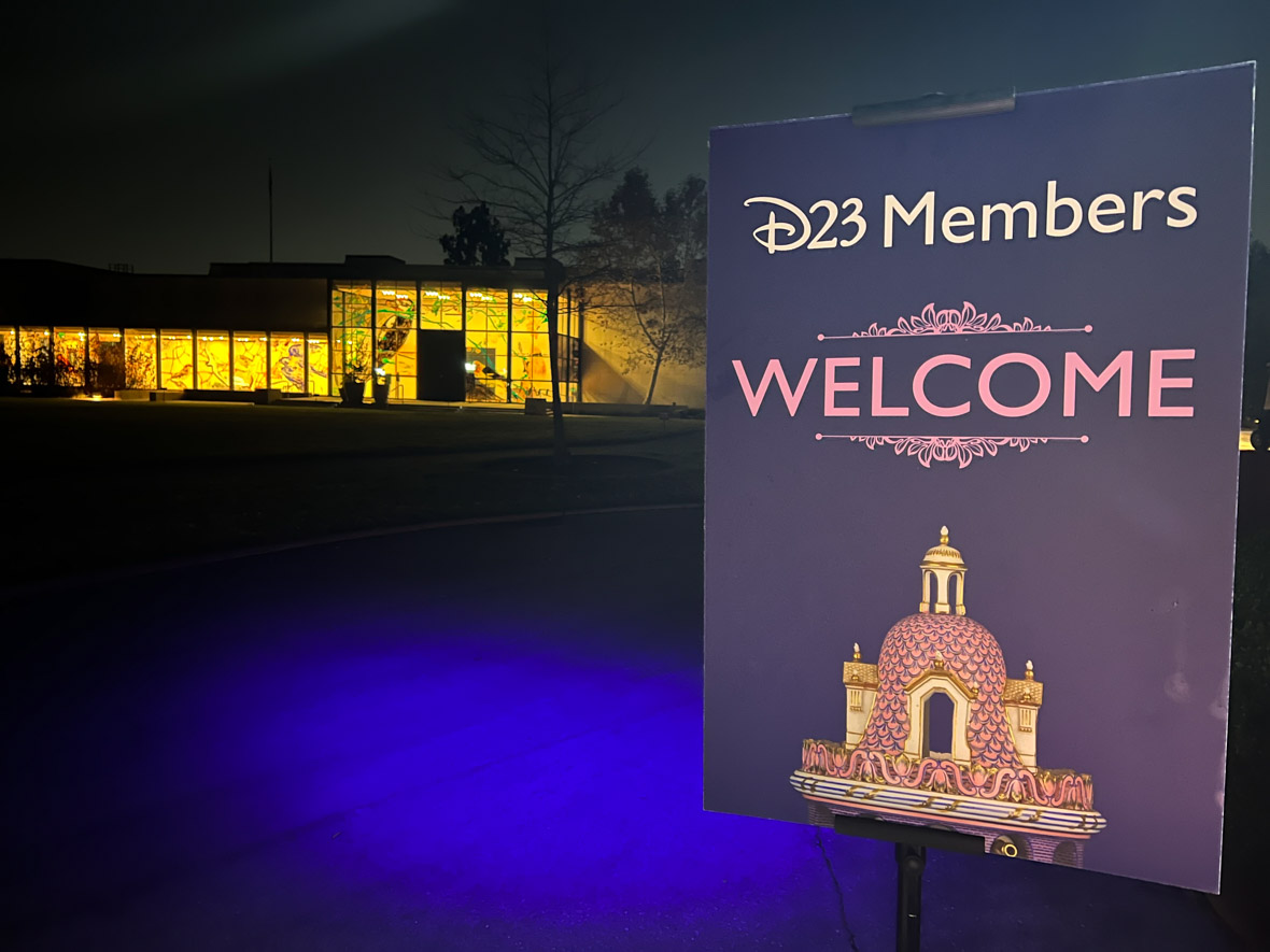 Welcome sign. The sign is purple with D23 Members written in white at the top. Below says Welcome in pink. Below is the top of the exhibit logo’s vase in pink.
