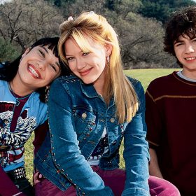 Miranda, Lizzie, and Gordo sit together in a grassy field. Miranda is smiling with her head on Lizzie’s shoulder. She has on a graphic tank top over a maroon long-sleeved shirt. Lizzie is sporting a closed-mouth smile and is wearing a studded jean jacket and maroon pants. Her blonde hair is styled half up and her bangs are parted in the middle. Gordo is standing behind the fence the girls are sitting on, smiling awkwardly at the camera, with a maroon T-shirt on.
