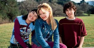 Miranda, Lizzie, and Gordo sit together in a grassy field. Miranda is smiling with her head on Lizzie’s shoulder. She has on a graphic tank top over a maroon long-sleeved shirt. Lizzie is sporting a closed-mouth smile and is wearing a studded jean jacket and maroon pants. Her blonde hair is styled half up and her bangs are parted in the middle. Gordo is standing behind the fence the girls are sitting on, smiling awkwardly at the camera, with a maroon T-shirt on.