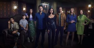 From left to right, actors Lex Lumpkin, Henry Joseph Samiri, Aliyah Royale, Warren Christie, Mariel Molino, Jon Ecker, Amy Acker, and Kelly Bishop pose in a promotional photo for Freeform’s The Watchful Eye. No one is smiling. They are standing in a dimly lit room with ornate wallpaper and portraits hanging on the walls.