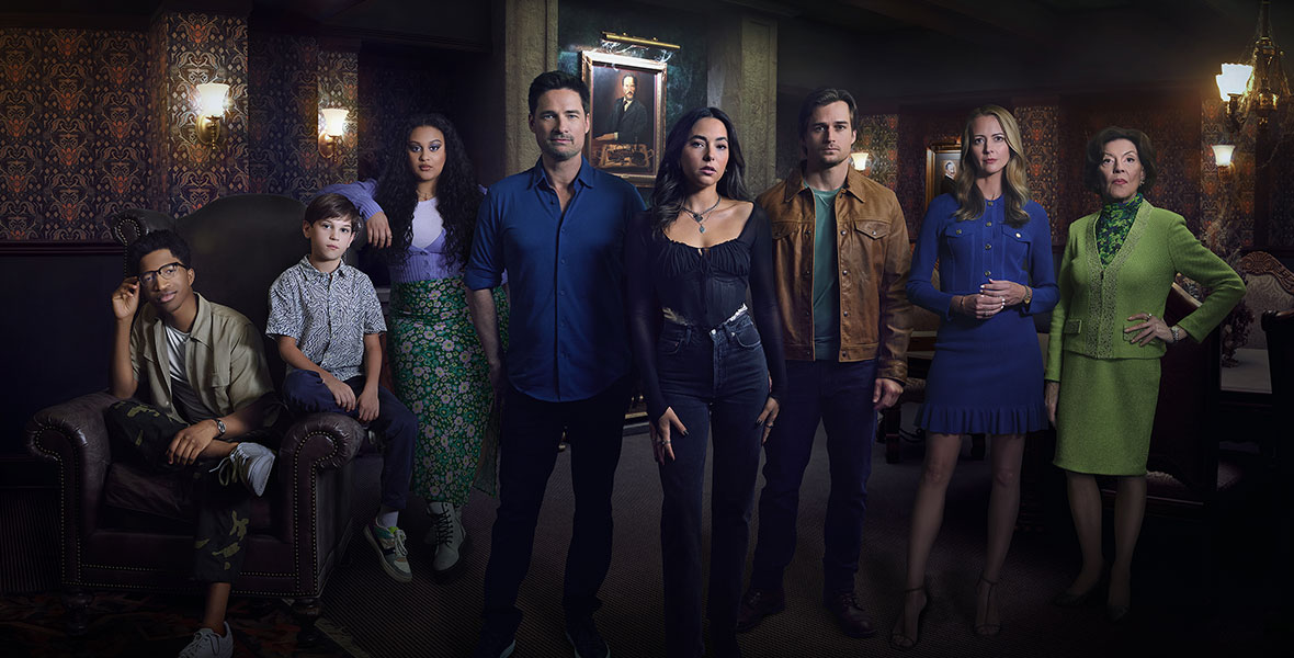 From left to right, actors Lex Lumpkin, Henry Joseph Samiri, Aliyah Royale, Warren Christie, Mariel Molino, Jon Ecker, Amy Acker, and Kelly Bishop pose in a promotional photo for Freeform’s The Watchful Eye. No one is smiling. They are standing in a dimly lit room with ornate wallpaper and portraits hanging on the walls.