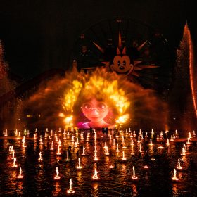 In an image of World of Color – One at Disney California Adventure Park, Mirabel from Encanto can be seen projected onto a screen of water—which is surrounded by hundreds of other fountains, both tall and short, that are spouting water and lit up with orange and yellow lights. The Pixar Pier Ferris wheel can be seen slightly behind the screen of water.