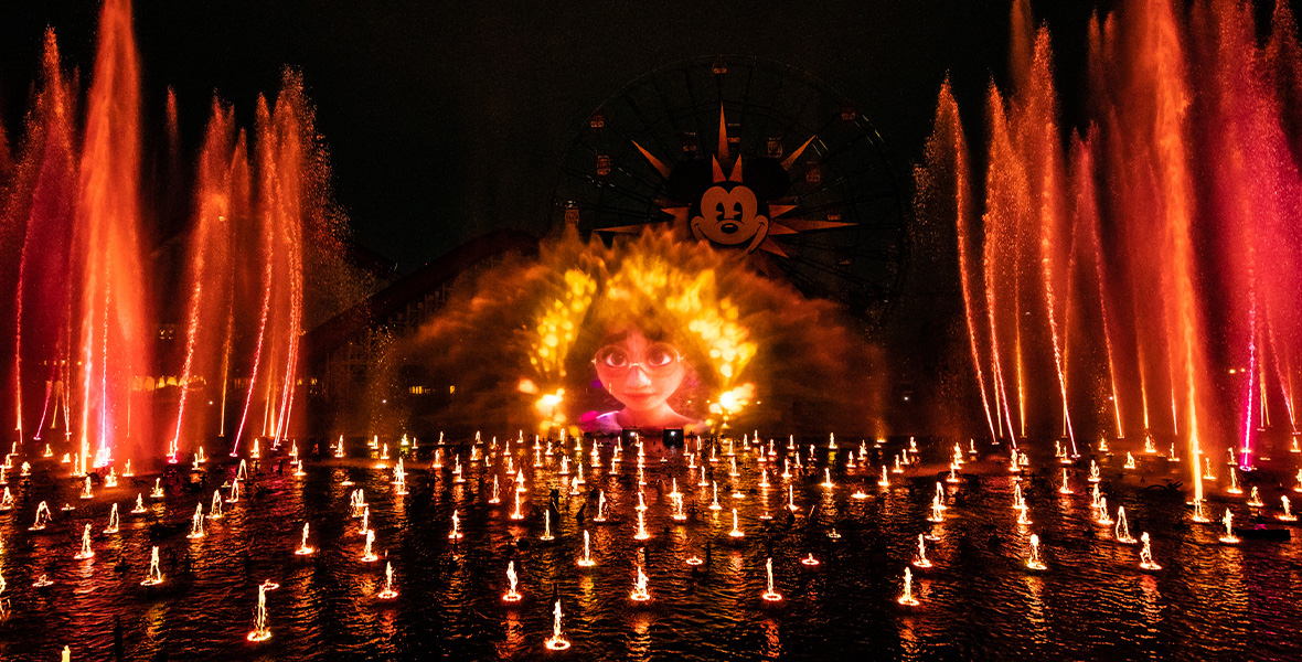 In an image of World of Color – One at Disney California Adventure Park, Mirabel from Encanto can be seen projected onto a screen of water—which is surrounded by hundreds of other fountains, both tall and short, that are spouting water and lit up with orange and yellow lights. The Pixar Pier Ferris wheel can be seen slightly behind the screen of water.