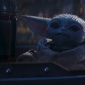 In an image from Lucasfilm’s The Mandalorian, Grogu is seen in the middle of the image, with the Mandalorian to his left. They appear to be sitting in the cockpit of a space vehicle; a dome is seen over their heads, and some buttons and lights are seen behind Grogu to the right.