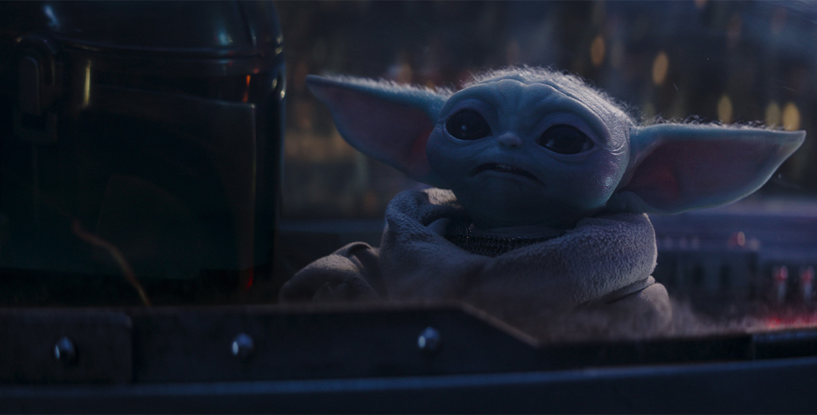 In an image from Lucasfilm’s The Mandalorian, Grogu is seen in the middle of the image, with the Mandalorian to his left. They appear to be sitting in the cockpit of a space vehicle; a dome is seen over their heads, and some buttons and lights are seen behind Grogu to the right.