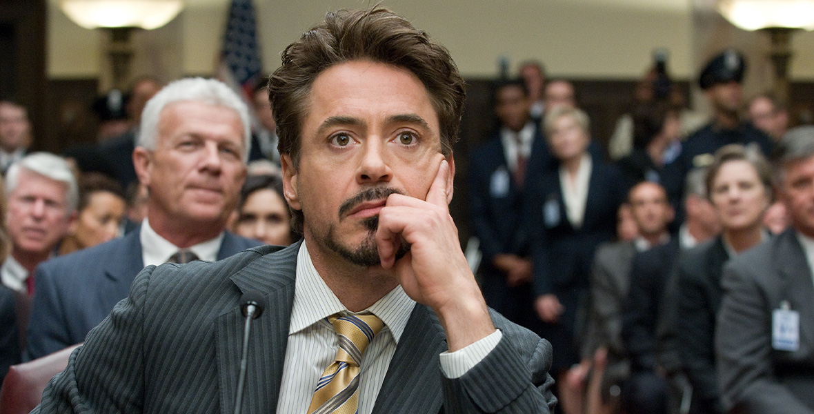 In a scene from Marvel Studios’ Iron Man 2, actor and Disney Legend Robert Downey Jr. portrays Tony Stark/Iron Man. He sits in a packed courtroom and behind a tiny microphone. He wears a dark gray pinstripe suit, a tan button-down shirt, and a yellow and gray striped tie.
