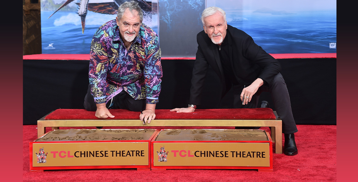 Jon Landau (left) and James Cameron (right) are seen bending down and leaning on a long red carpeted footstool above two boxes full of cement that are both labeled with signs reading “TCL Chinese Theatre.” A poster for “Avatar: The Way of Water” is seen behind them. Landau is wearing a print button-up shirt; Cameron is wearing a dark suit. Both are smiling and looking toward the camera.