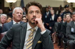 In a scene from Marvel Studios’ Iron Man 2, actor and Disney Legend Robert Downey Jr. portrays Tony Stark/Iron Man. He sits in a packed courtroom and behind a tiny microphone. He wears a dark gray pinstripe suit, a tan button-down shirt, and a yellow and gray striped tie.