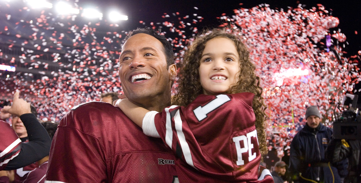 In a scene from the feature film The Game Plan, actor Dwayne Johnson portrays Joe Kingman and holds actor Madison Pettis, who plays Peyton, a young girl, in his arms. Johnson and Pettis both wear maroon football jerseys with white and black stripes and lettering. Red and white confetti falls from the sky.