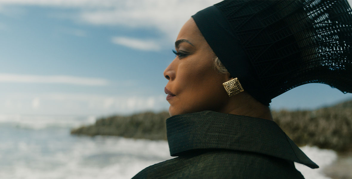 Angela Bassett wears an ornate black headpiece; a large square, gold earring; and a black cloak. She stands along the coast and stares off into the distance.