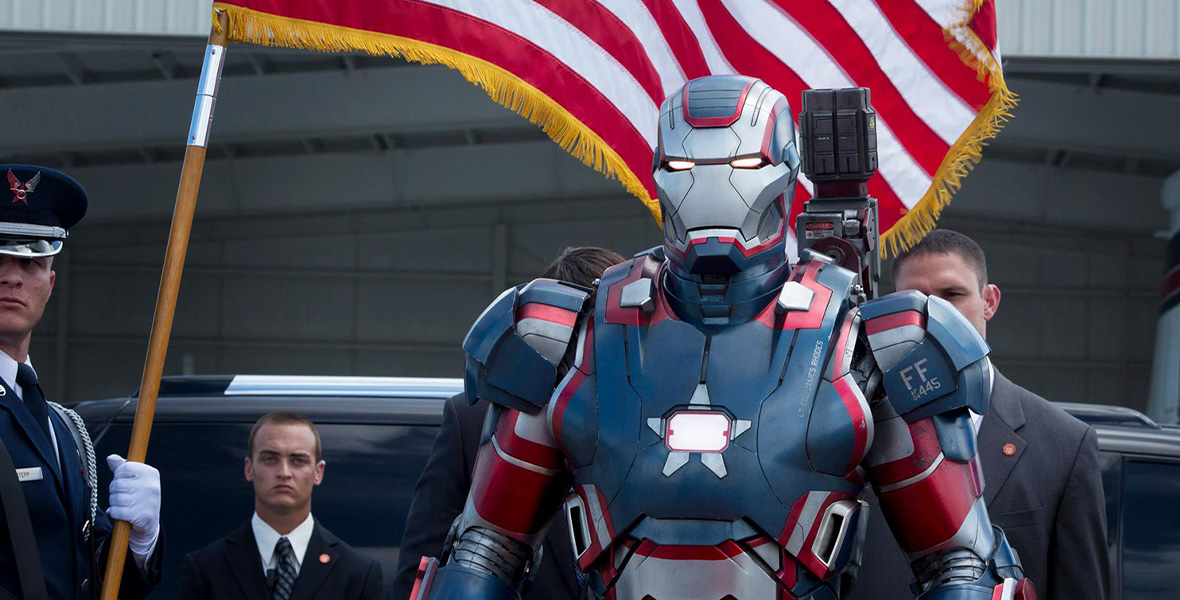 In a scene from Marvel Studios’ Iron Man 3, a man wears a red, white, and blue suit of armor and walks next to a man holding an American flag.