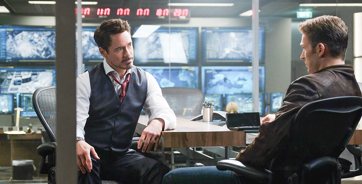 In a scene from Marvel Studios’ Captain America: Civil War, actor and Disney Legend Robert Downey Jr. as Tony Stark/Iron Man sits at a wooden conference table and looks at actor Chris Evans as Steve Rogers/Captain America. Behind them are large TV screens displaying surveillance footage and clocks displaying various time zones.