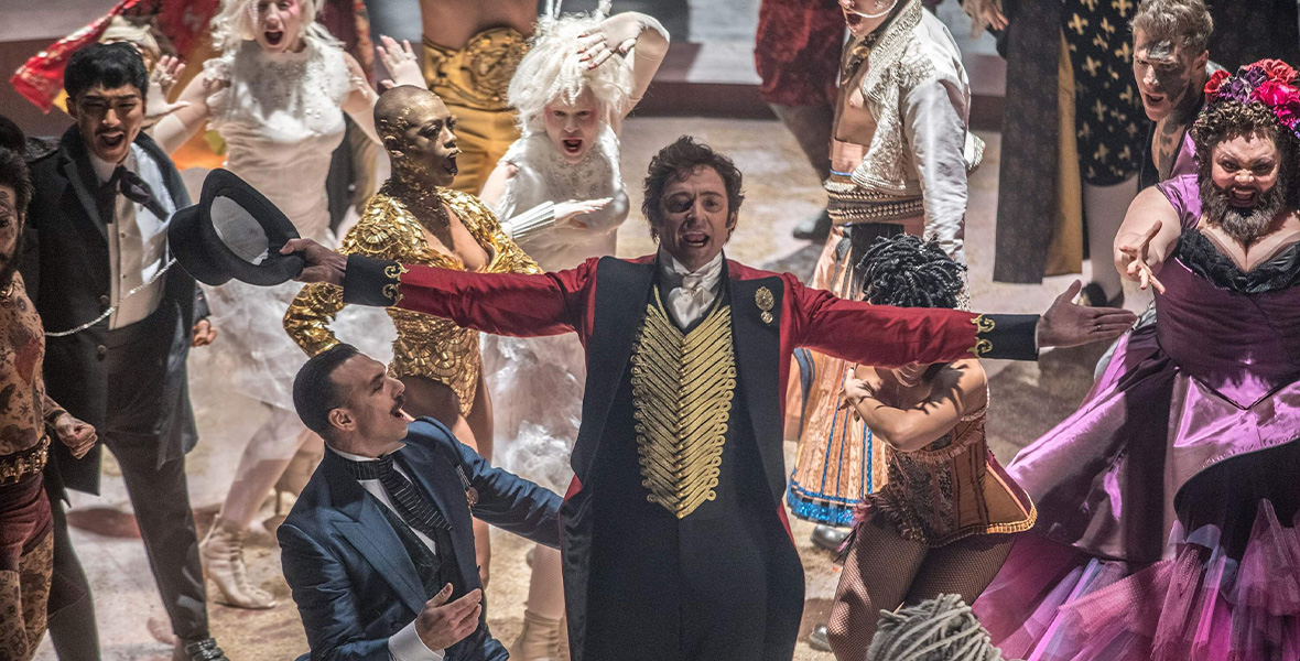 In a scene from the film The Greatest Showman, actor Hugh Jackman portrays P.T. Barnum and wears a ringmaster outfit consisting of a long red jacket with black lapels, a gold vest, a white, collared shirt, and black pants. Jackman is surrounded by a crowd of people all dressed in elaborate circus costumes.