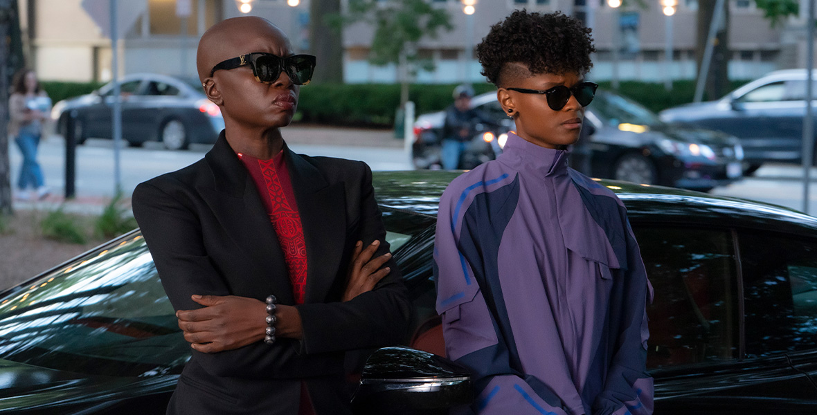In a scene from Black Panther: Wakanda Forever, actors Danai Gurira as Okoye and Letitia Wright as Shuri stand next to each other and lean against a black sedan. Gurira wears black sunglasses, a black blazer, and a red dress and folds her arms across her abdomen. Wright wears black sunglasses and a two-piece purple tracksuit with dark purple and electric blue color blocking.