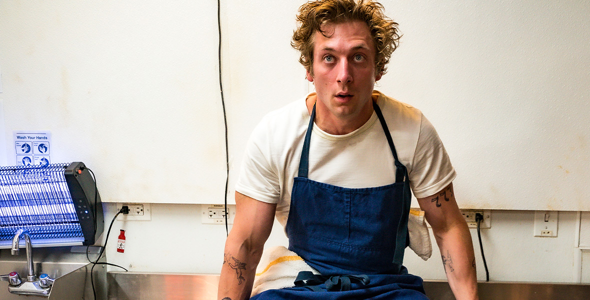 Jeremy Allen White sits on a somewhat dirty steel countertop in an industrial kitchen. He is wearing a white T-shirt, exposing multiple tattoos on each arm. The tattoos include Cupid, a snail, slot machine numbers, and more. White is wearing a blue apron over his shirt. His curly blond hair is messy, and he appears exasperated.