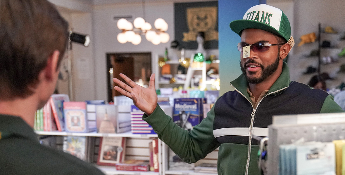 In a scene from Freeform series grown-ish, Trevor Jackson portrays Aaron and stands in the Cal U bookstore. He wears sunglasses, a hat, and jacket, all with tags hanging off them.