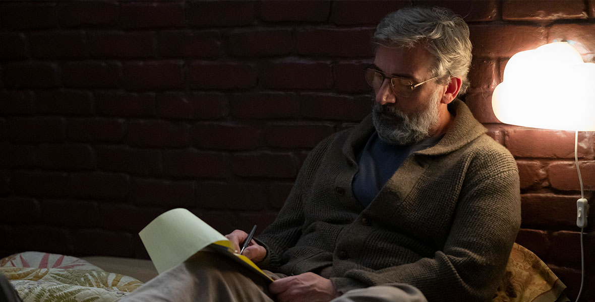 Steve Carell sits in bed against a brick wall. To the left of him is a large lamp. Carell’s hair is white and his beard is white and full. He is wearing eyeglasses, a chunky green sweater, khaki pants, and brown dress shoes. He is writing on a yellow legal pad.