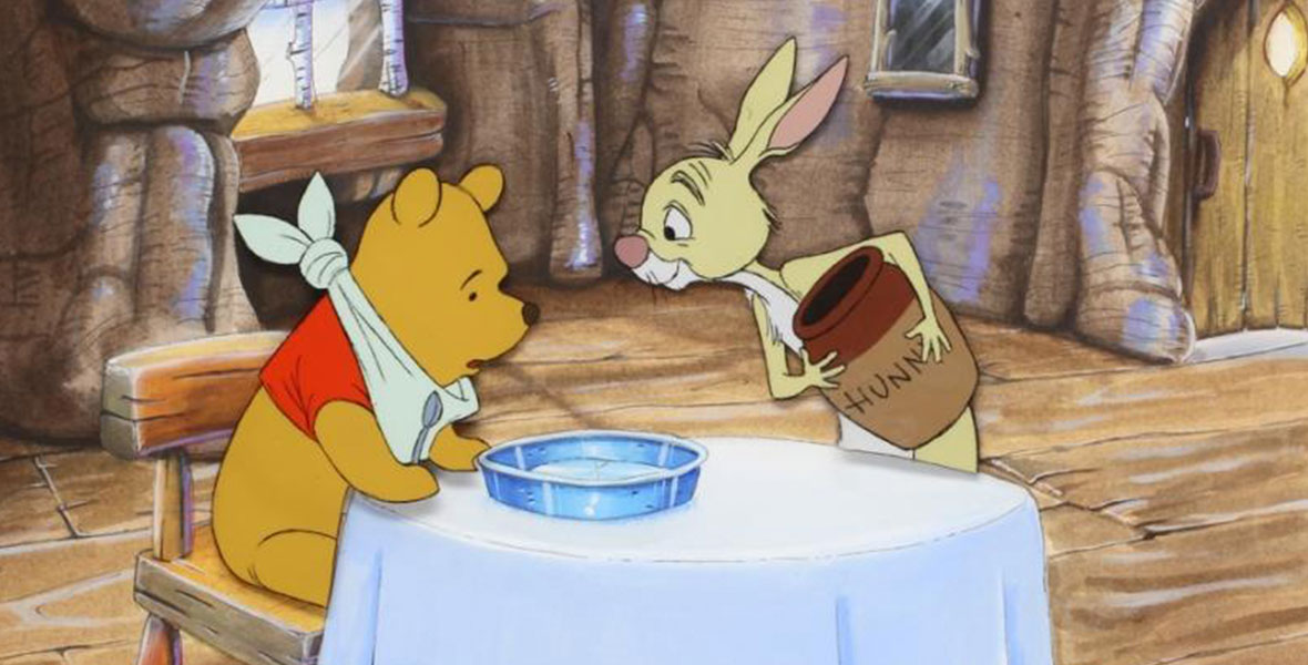 Winnie the Pooh sits at a table with a napkin around his neck as Rabbit stands across from him. Rabbit smiles and holds a jar that reads “Hunny” as Pooh holds a spoon in front of an empty plate.