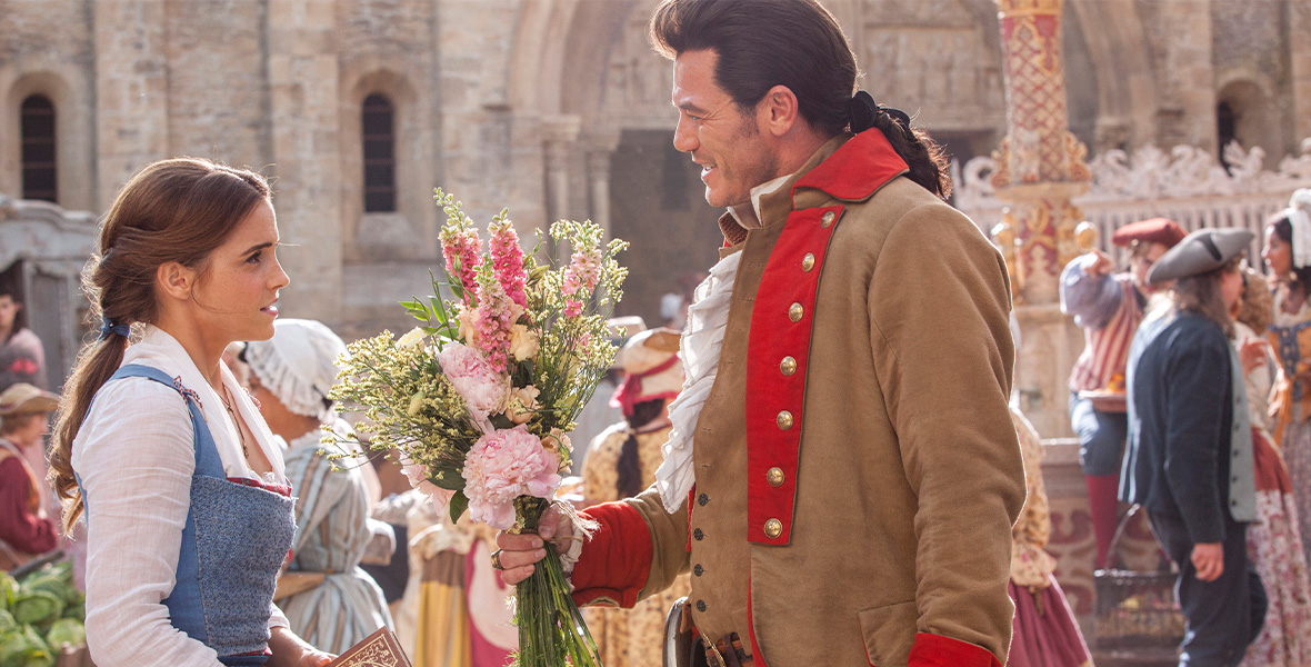 In a scene from Beauty and the Beast, actors Emma Watson as Belle and Luke Evans as Gaston stand in a town square. Watson wears a white, long-sleeved blouse and a light blue dress while she holds a book with a brown cover in her hands. Evans holds a bouquet of pink peonies and pink hyacinths in his right hand and wears a tan coat with a red lapel and a red collar.