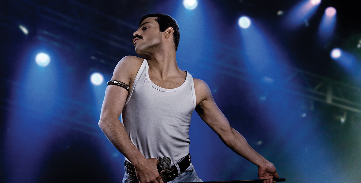In a scene from the film Bohemian Rhapsody, actor Rami Malek portrays singer Freddie Mercury. He wears a black armband, a white tank top, blue denim jeans, and a black leather belt.