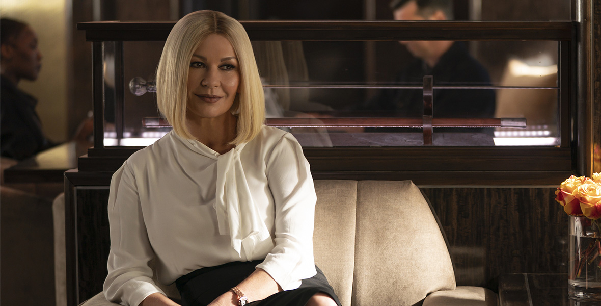 In a scene from National Treasure: Edge of History, actor Catherine Zeta-Jones portrays Billie and sits in a tan armchair. She wears a white blouse and a black skirt.