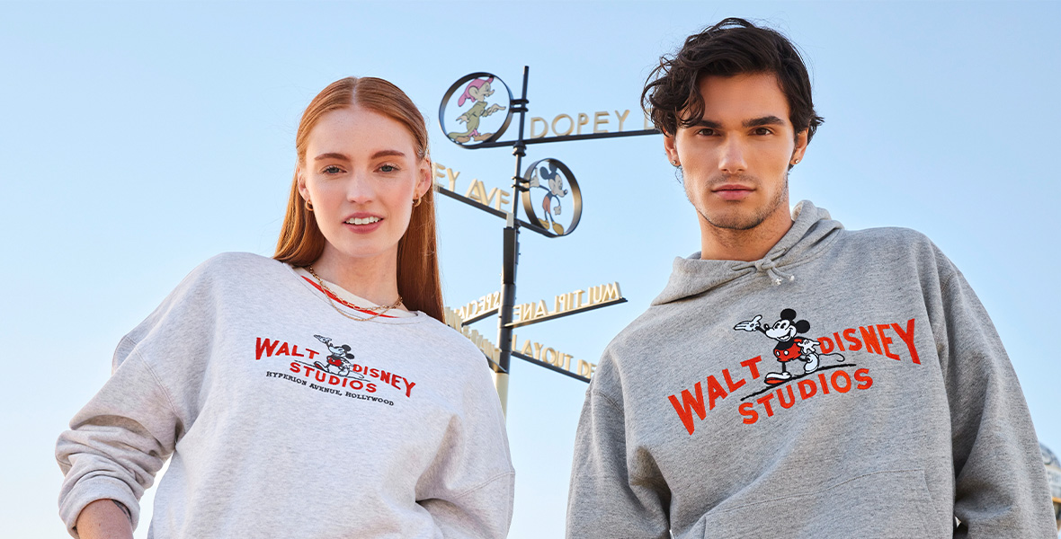 Two people stand side by side, with the blue sky behind them and the famous “street sign” from The Walt Disney Studios in Burbank, CA, between them, listing “Mickey Ave,” “Dopey Drive,” etc. The person on the left has long red hair and is wearing a lighter gray sweatshirt emblazoned with a vintage “Walt Disney Studios” logo in red; their left hand is in their jeans pocket. The person on the right has short brown hair and a scruffy beard and is wearing a darker gray hoodie emblazoned with a vintage “Walt Disney Studios” logo. 