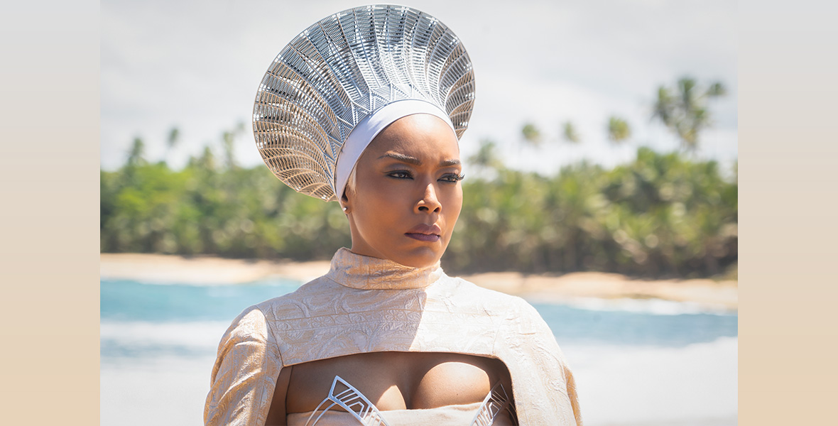 A close-up of Angela Bassett shows her wearing cream and white funeral attire. She stands on the coastline with trees in the background and the sun shining.